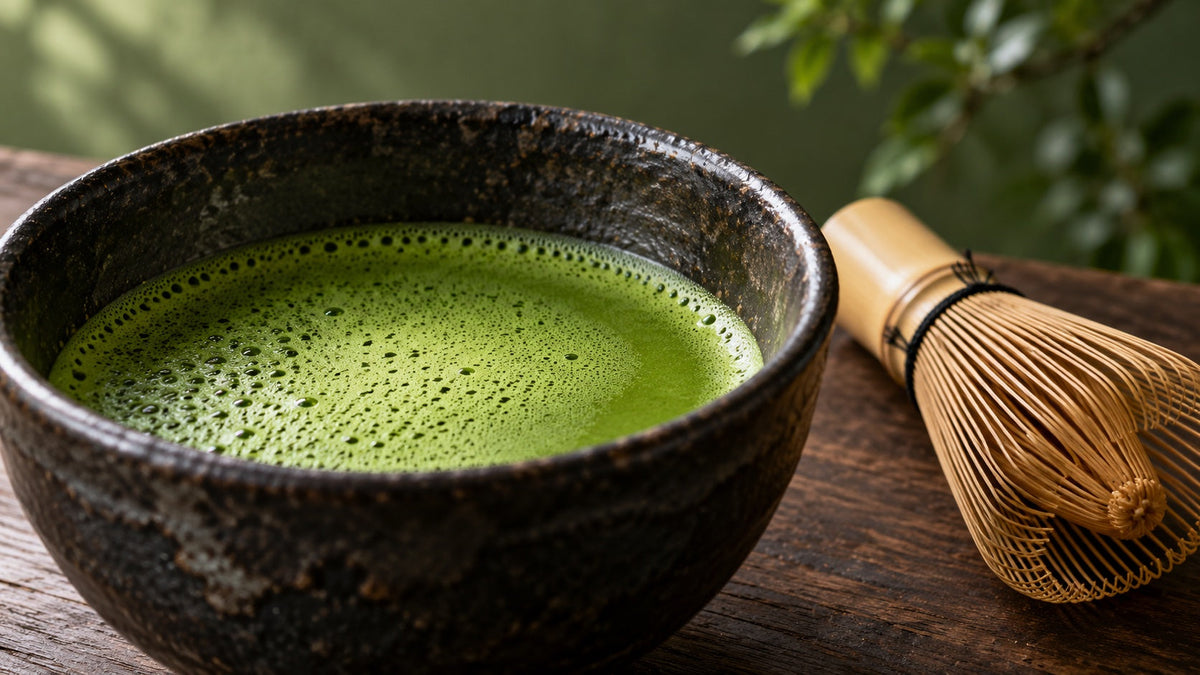 Feiner grüner Matcha-Schaum in einer dunklen Keramikschale neben einem Bambusbesen auf Walnussholz