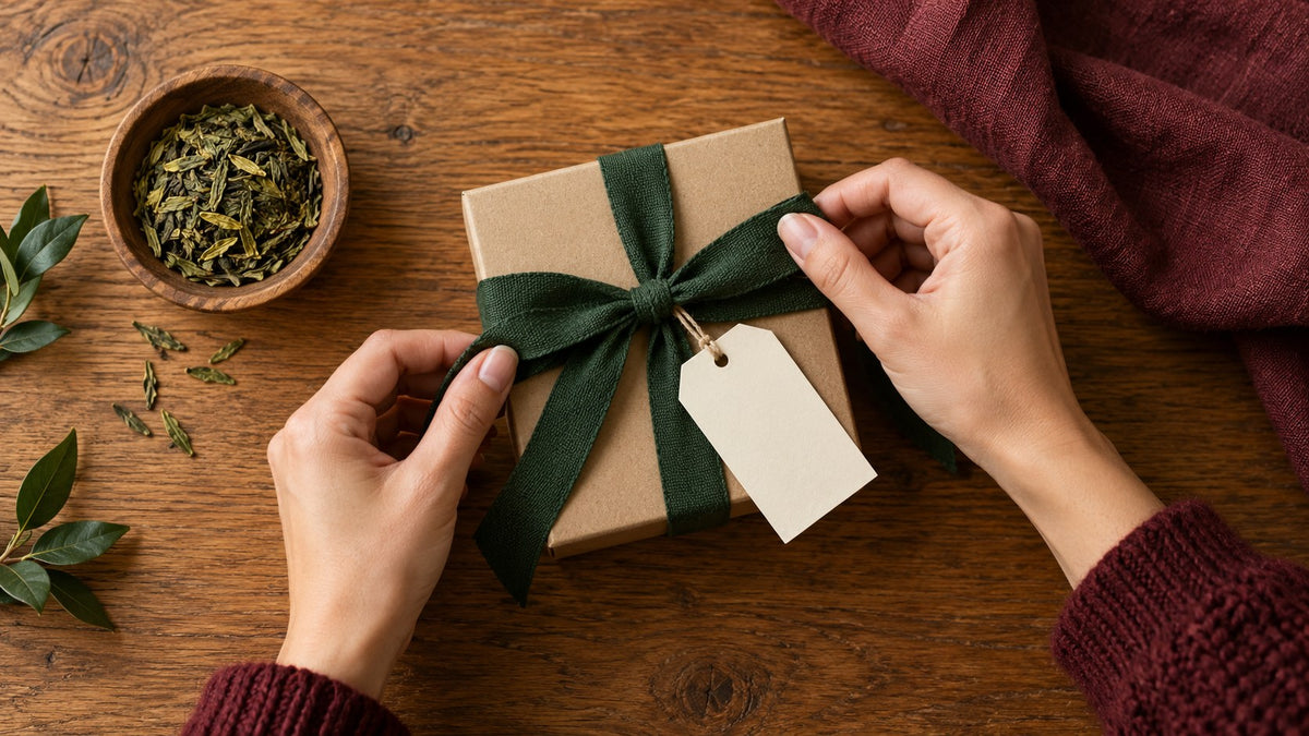 Hände binden eine dunkelgrüne Stoffschleife um eine geschlossene schlichte Geschenkbox auf einem warmen Holztisch, daneben ein leerer Anhänger und ein kleines Schälchen lose Teeblätter